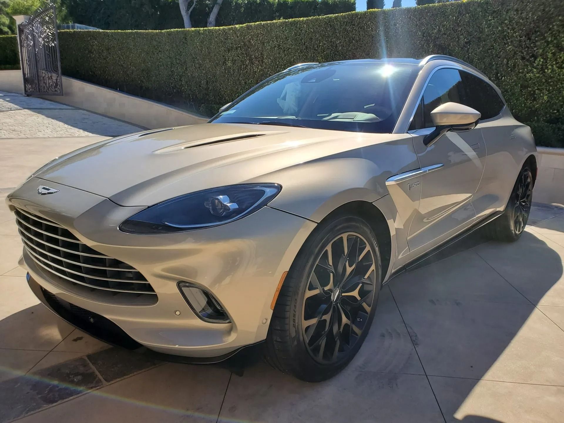 A metallic champagne-colored Aston Martin DBX SUV parked on a paved driveway in front of a hedge.