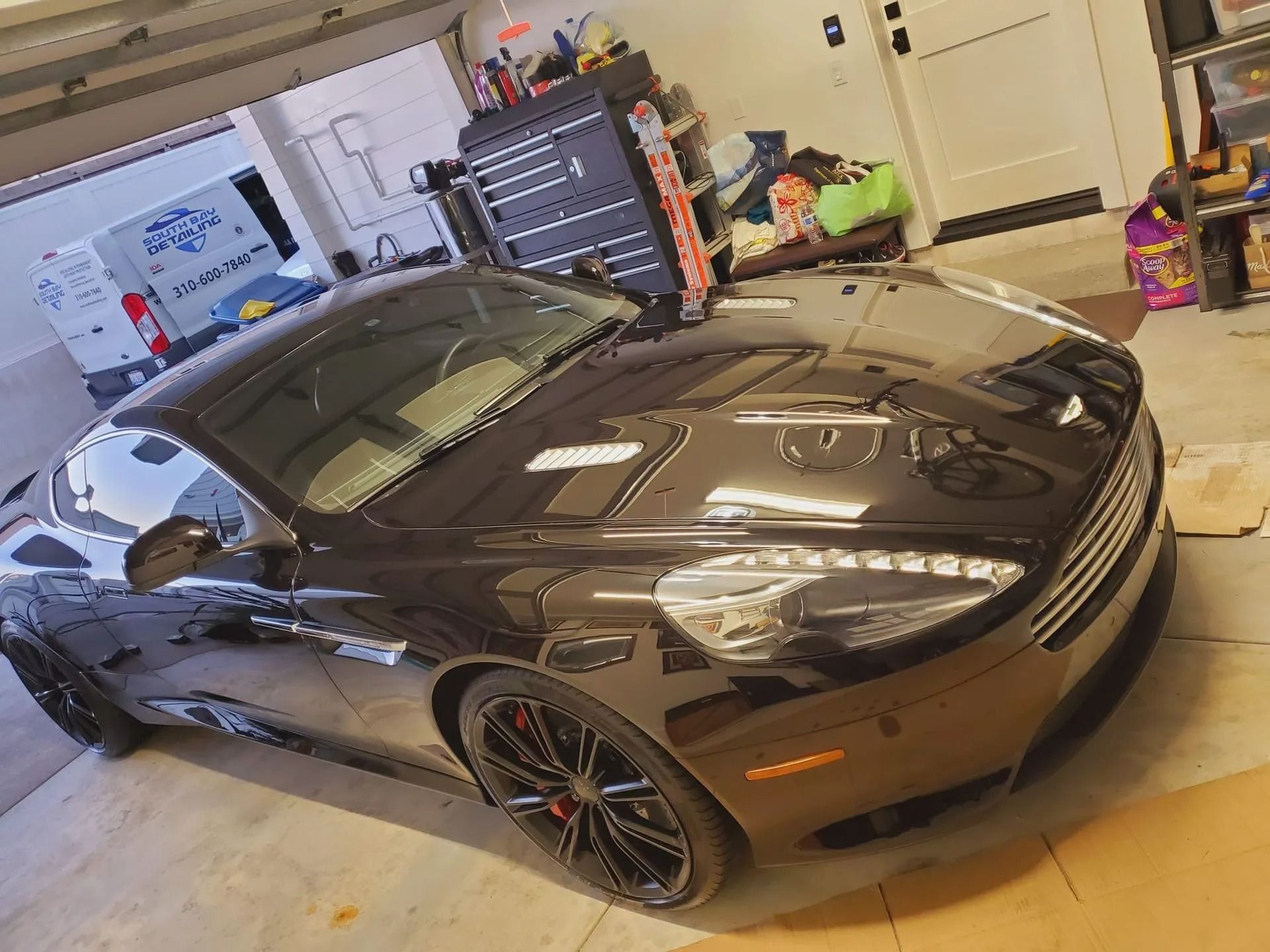 A black Aston Martin coupe parked inside a home garage, featuring alloy wheels and a sleek, shiny exterior finish.