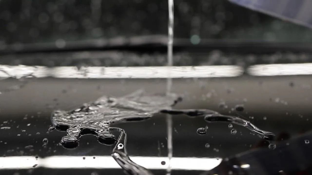 A thin stream of water pours onto a dark, reflective, slightly wet surface, creating small ripples and splashes.