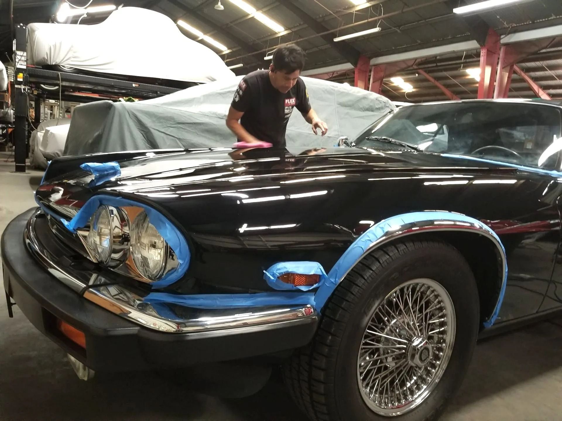 A person works on detailing the hood of a black Jaguar car inside a garage, with blue painter's tape on the trim.