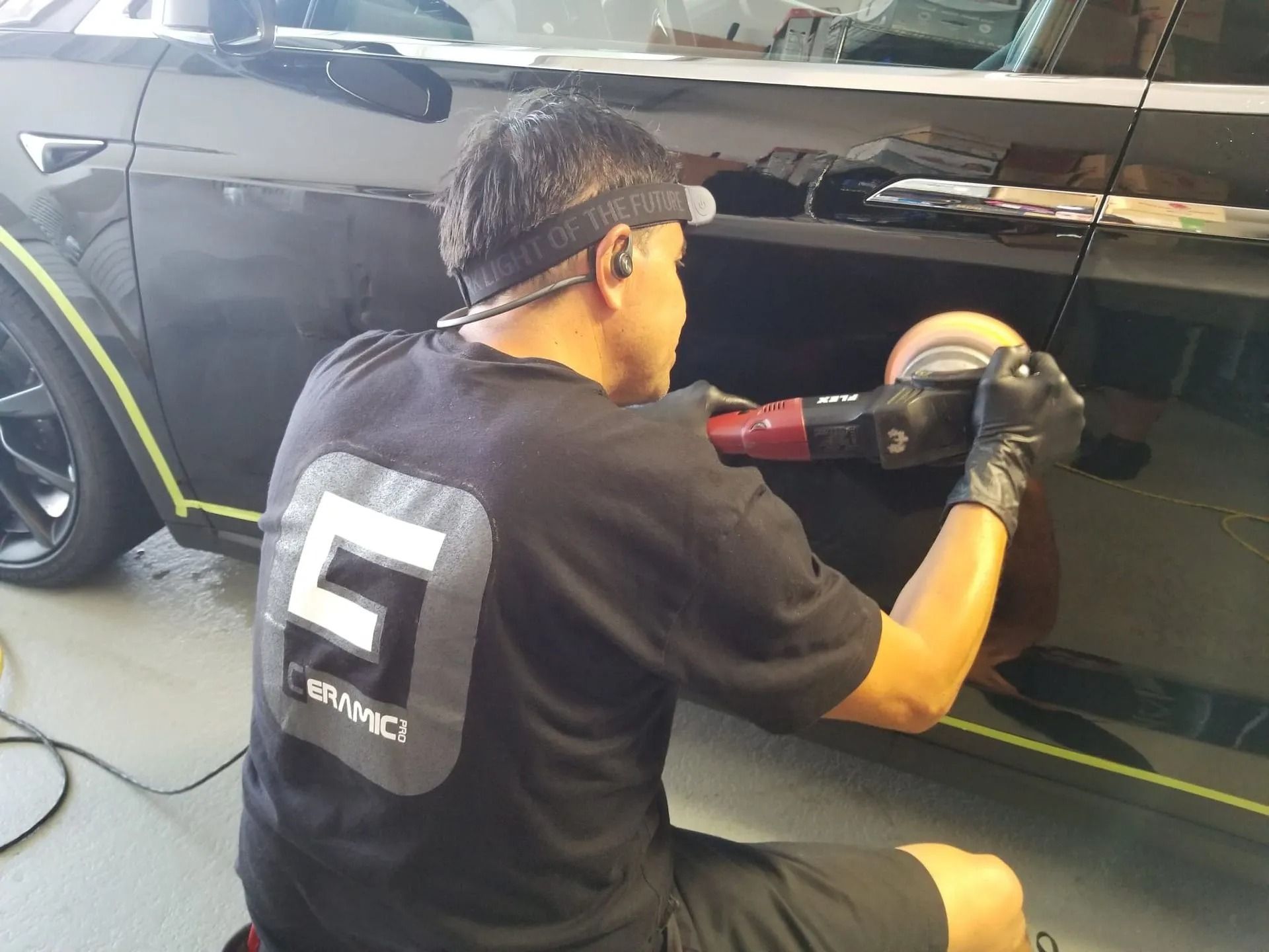 A person wearing a black shirt and headlamp uses a power buffer to polish the side of a black vehicle.
