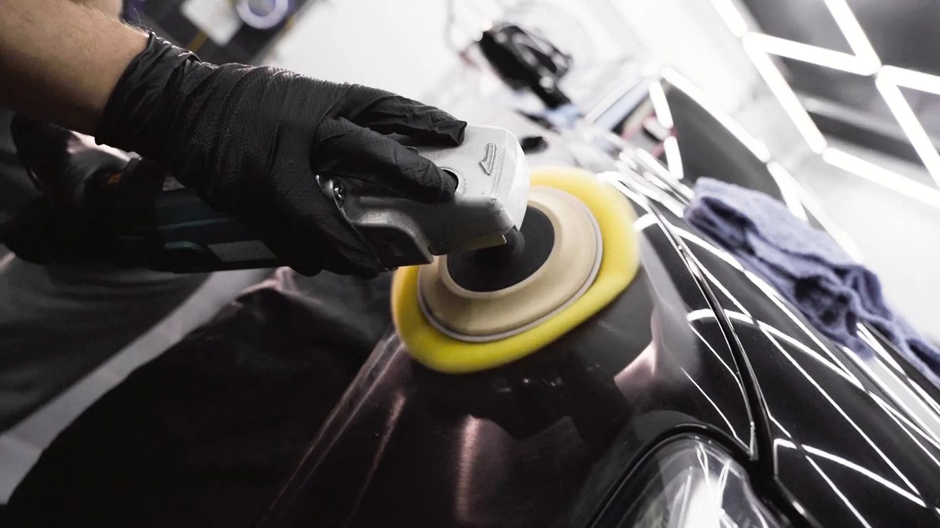A gloved hand uses a power buffer with a yellow pad to polish the black hood of a car in a workshop.