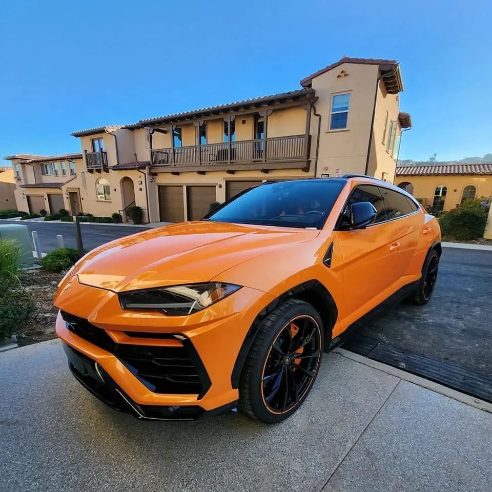 A bright orange Lamborghini Urus SUV parked on a paved driveway in front of a tan, multi-story stucco building.
