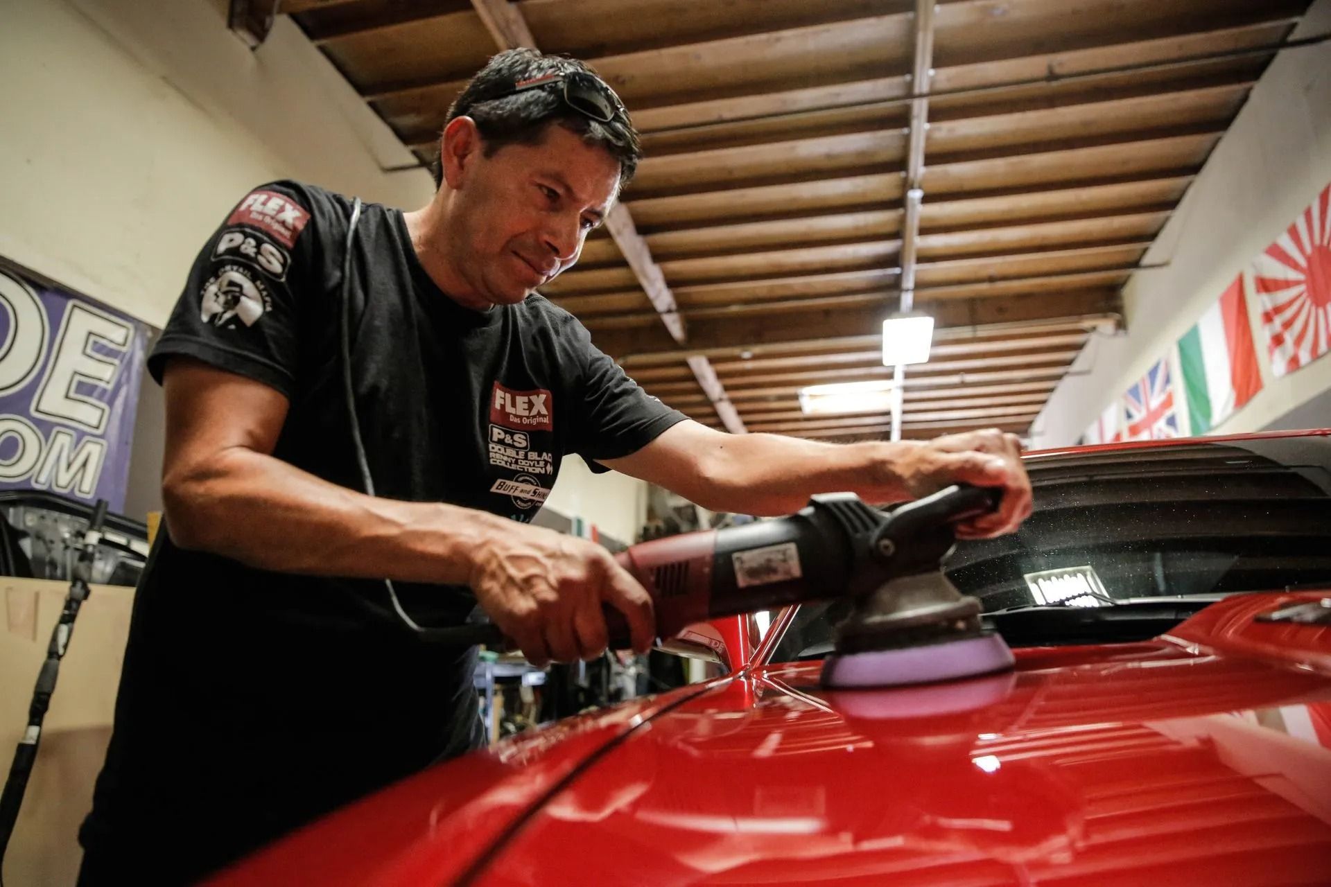 A technician uses a rotary polisher to buff the red hood of a car inside a garage workshop.