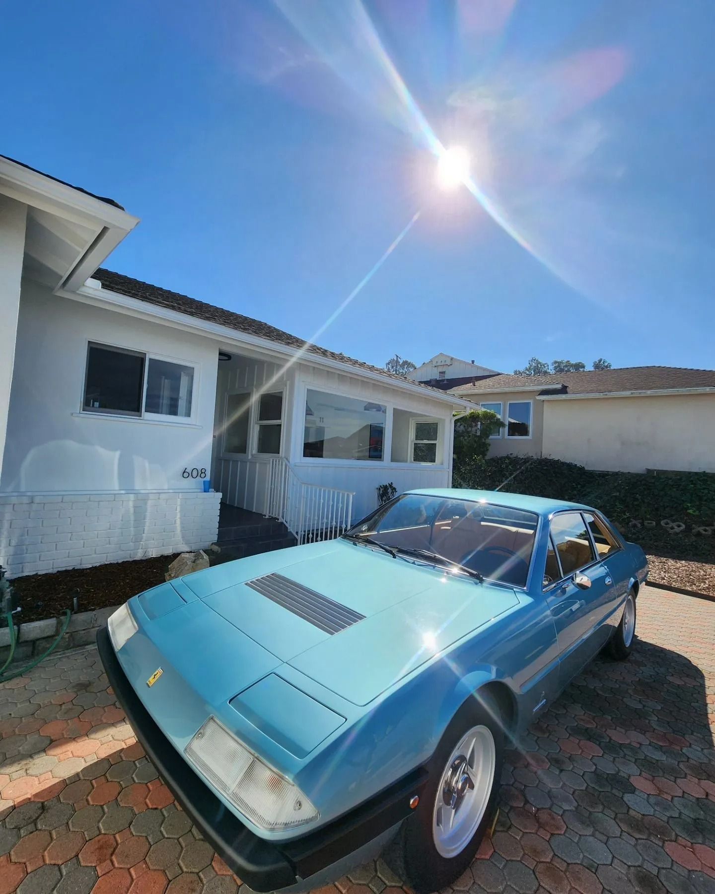 A light blue vintage sports car parked in a paved driveway in front of a white house on a bright, sunny day.