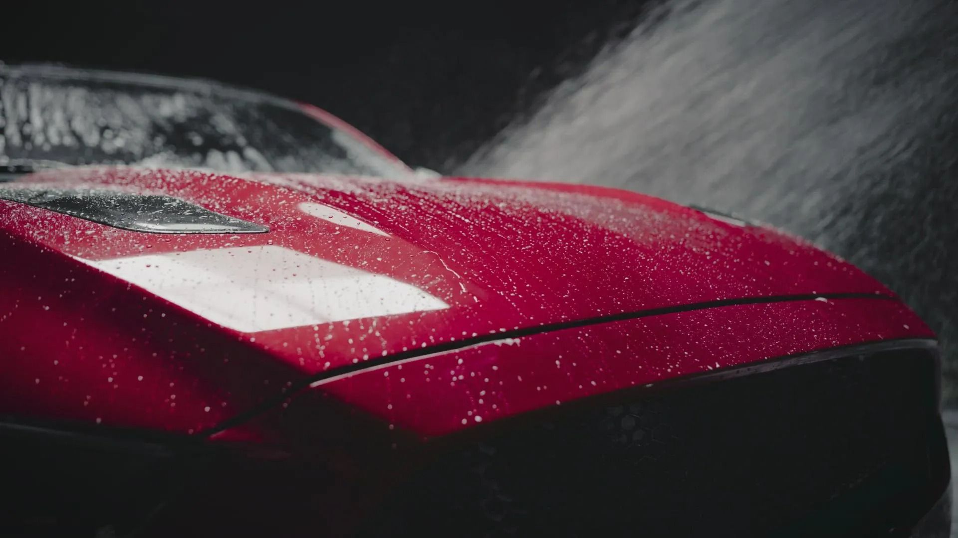 Water sprays across the shiny, wet hood of a red sports car in a dark setting.