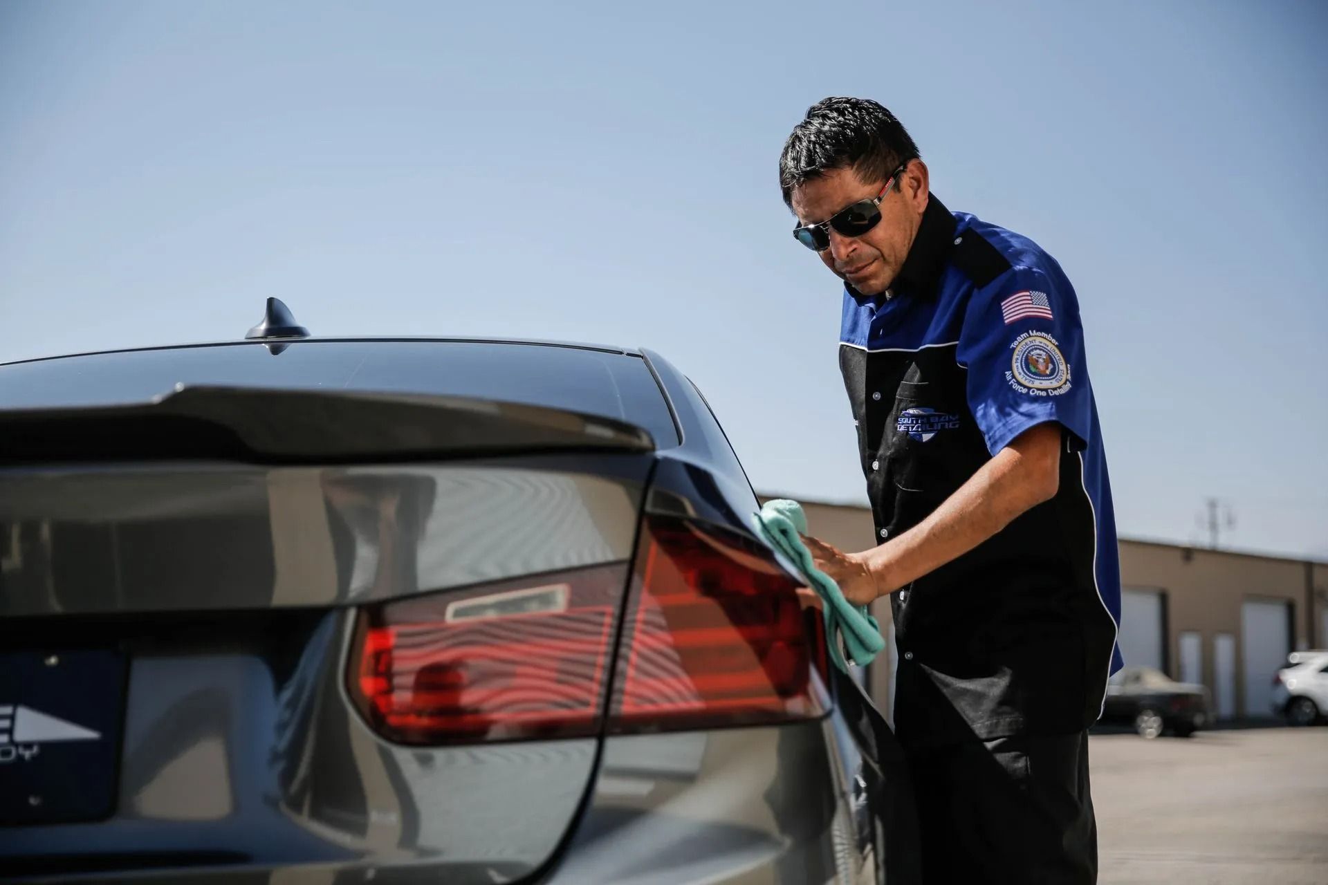 A person wearing sunglasses and a blue uniform shirt cleans the taillight of a grey car with a green cloth.