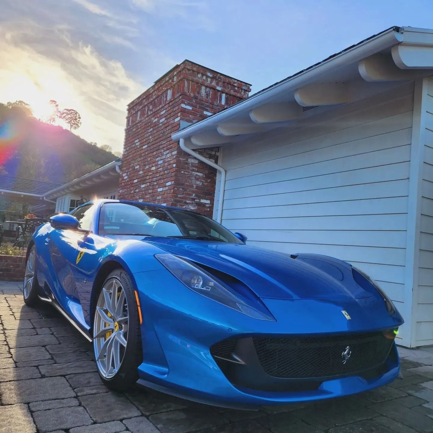 A metallic blue Ferrari 812 parked on a paved driveway next to a white building with a brick chimney under a sunny sky.