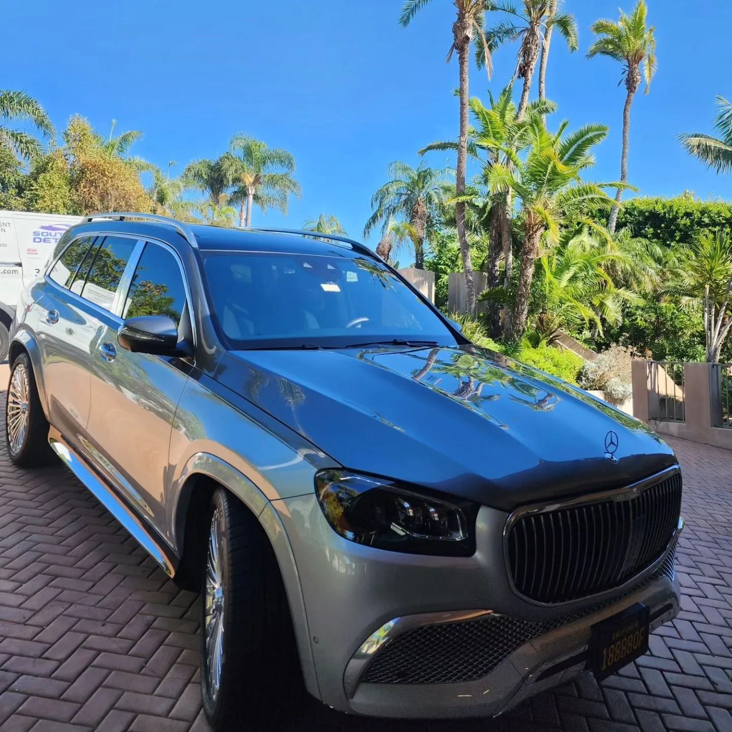 A silver Mercedes-Maybach GLS SUV parked on a paved driveway surrounded by palm trees under a clear blue sky.