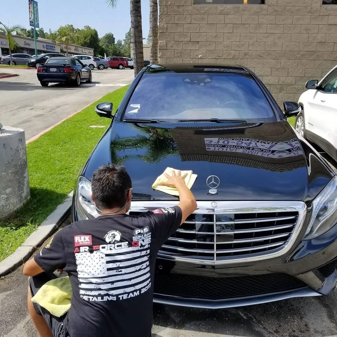 A person cleans the hood of a shiny black Mercedes-Benz parked in an outdoor lot.