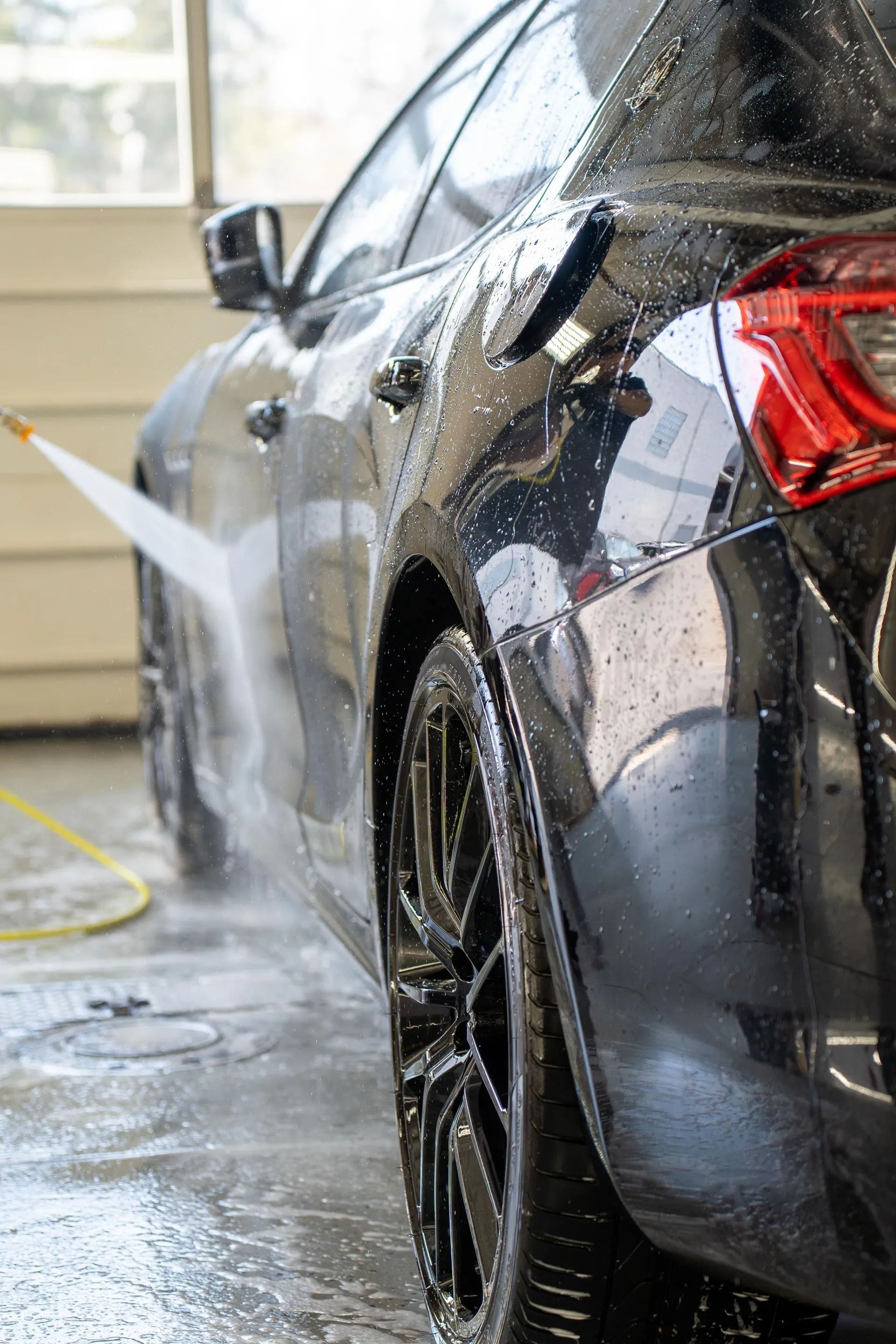 A person sprays water from a pressure washer onto the side of a black car parked in a garage.