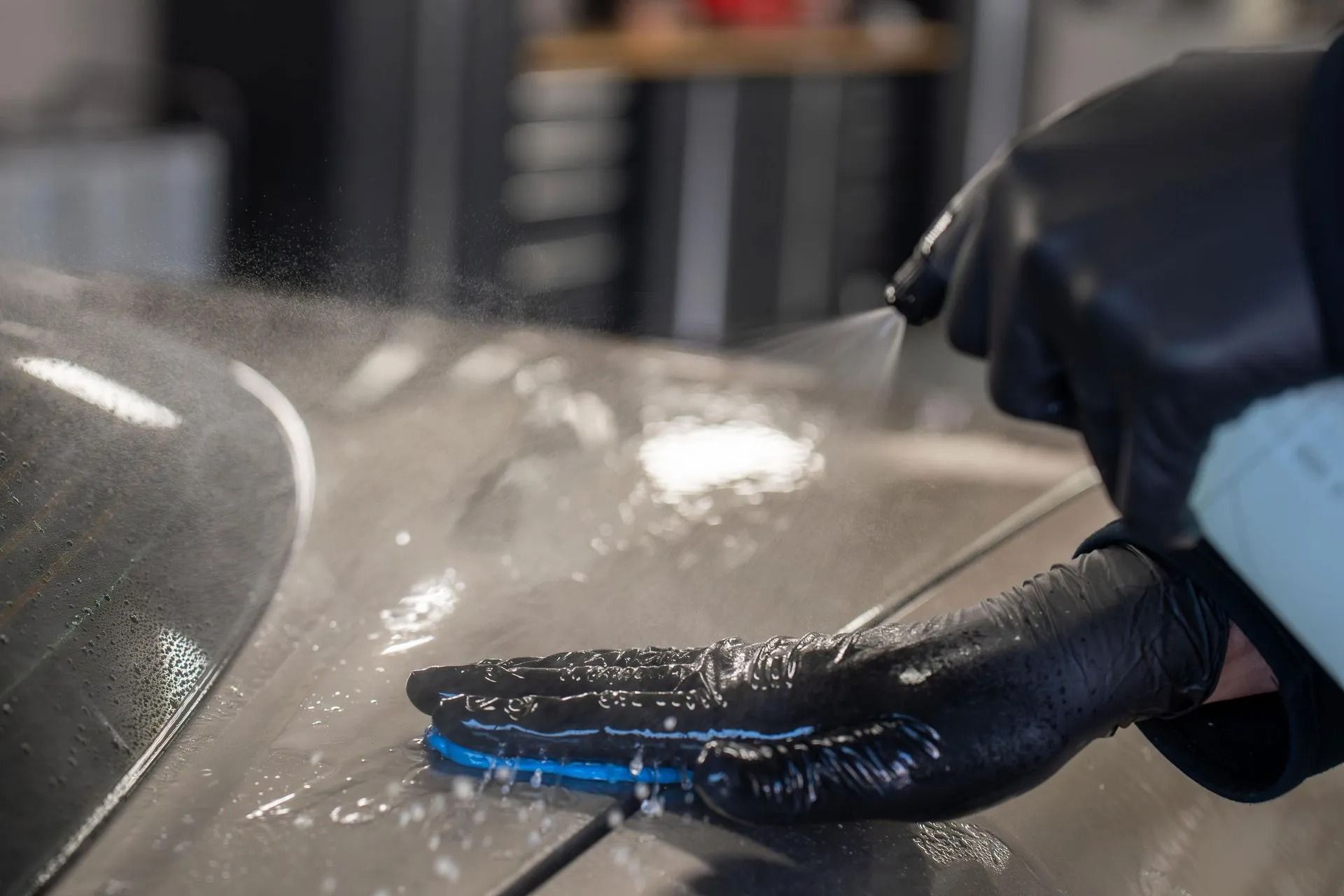 A person wearing black gloves sprays liquid onto a car surface while using a blue applicator pad to spread it.