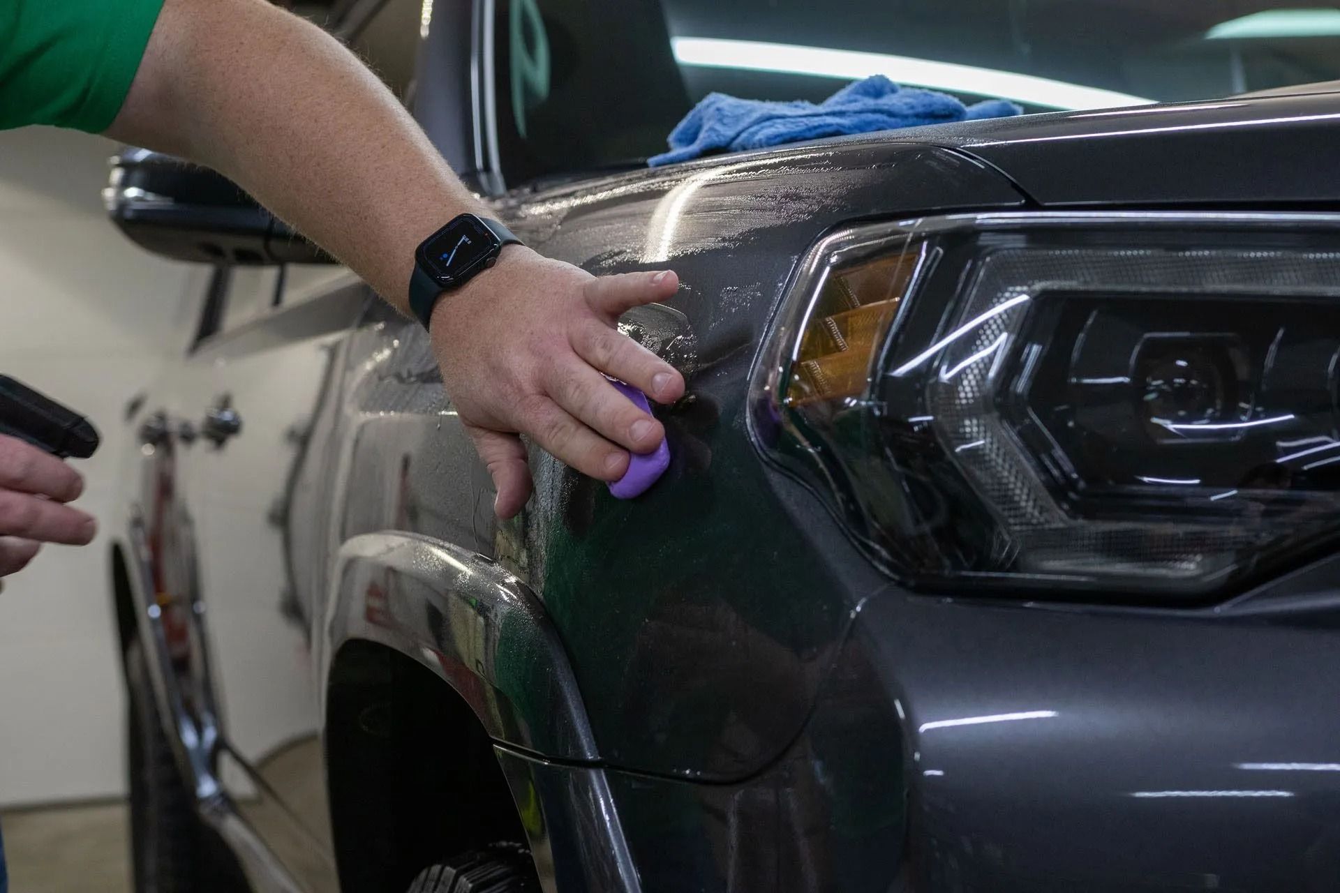 A person uses a purple clay bar to clean the dark gray paint on the front fender of a vehicle near the headlight.