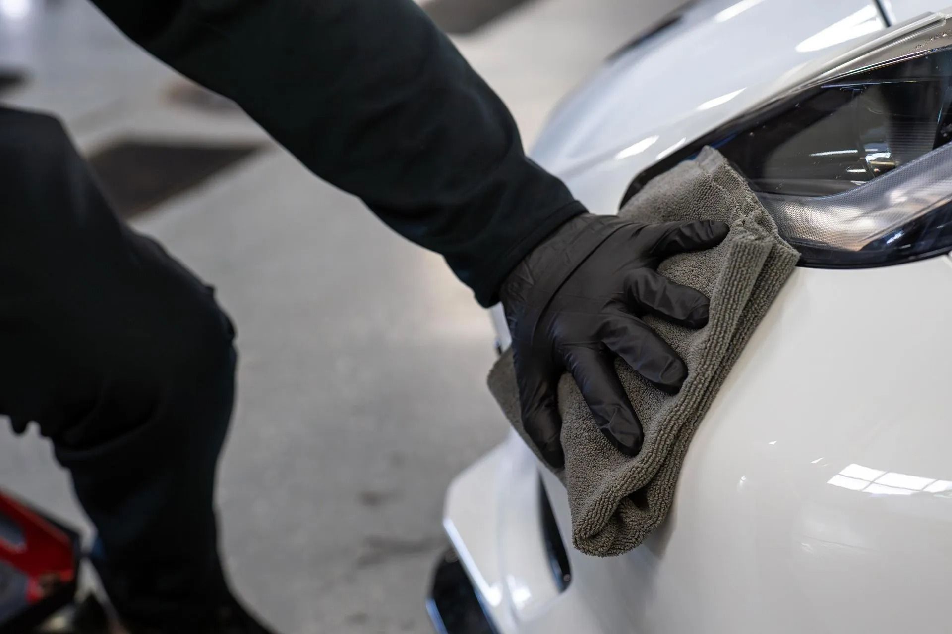 A person wearing black gloves wipes a white car with a gray microfiber cloth in a workshop.
