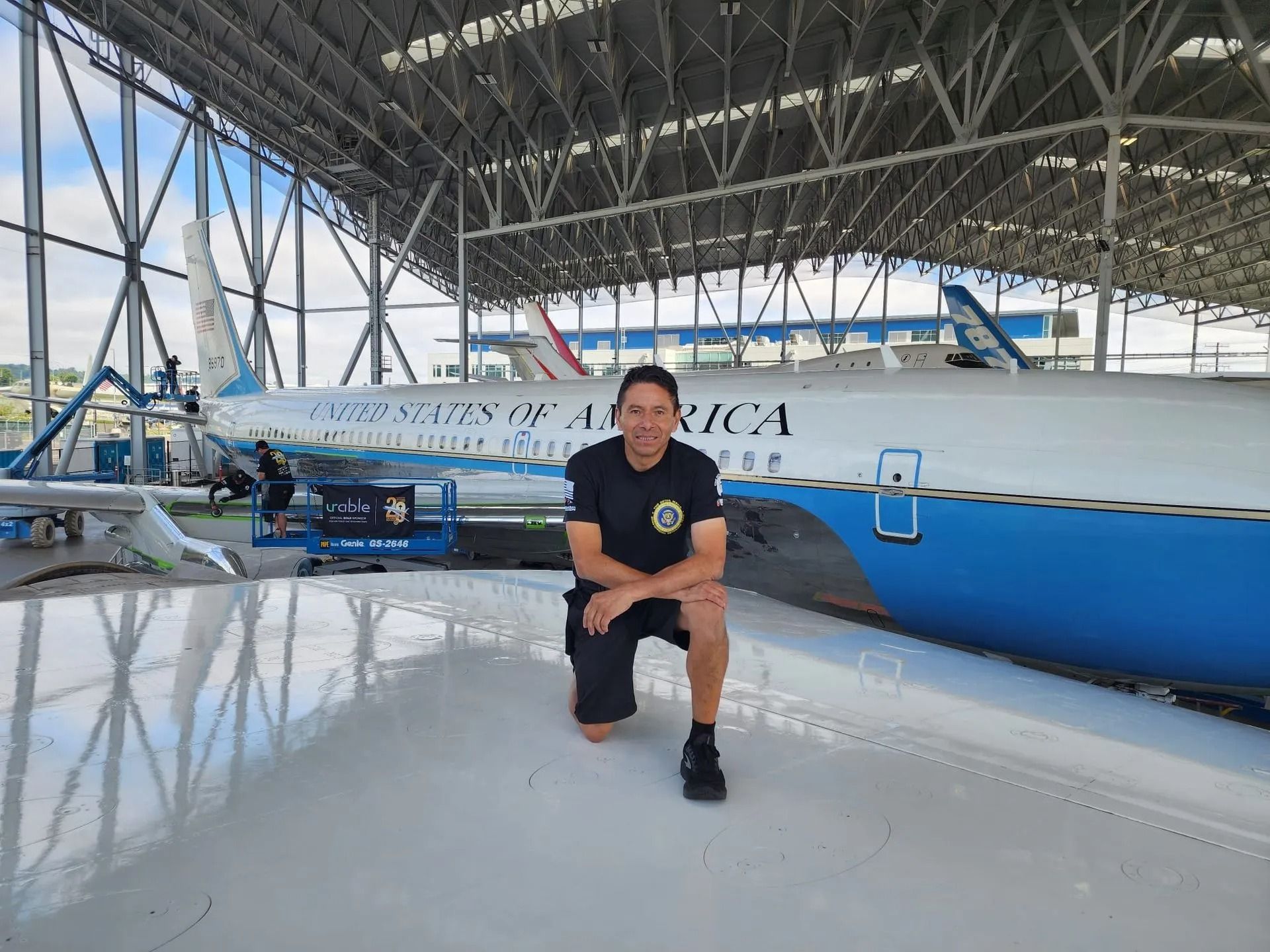 A person in a dark t-shirt kneels on an airplane wing inside a hangar, with a United States of America aircraft nearby.