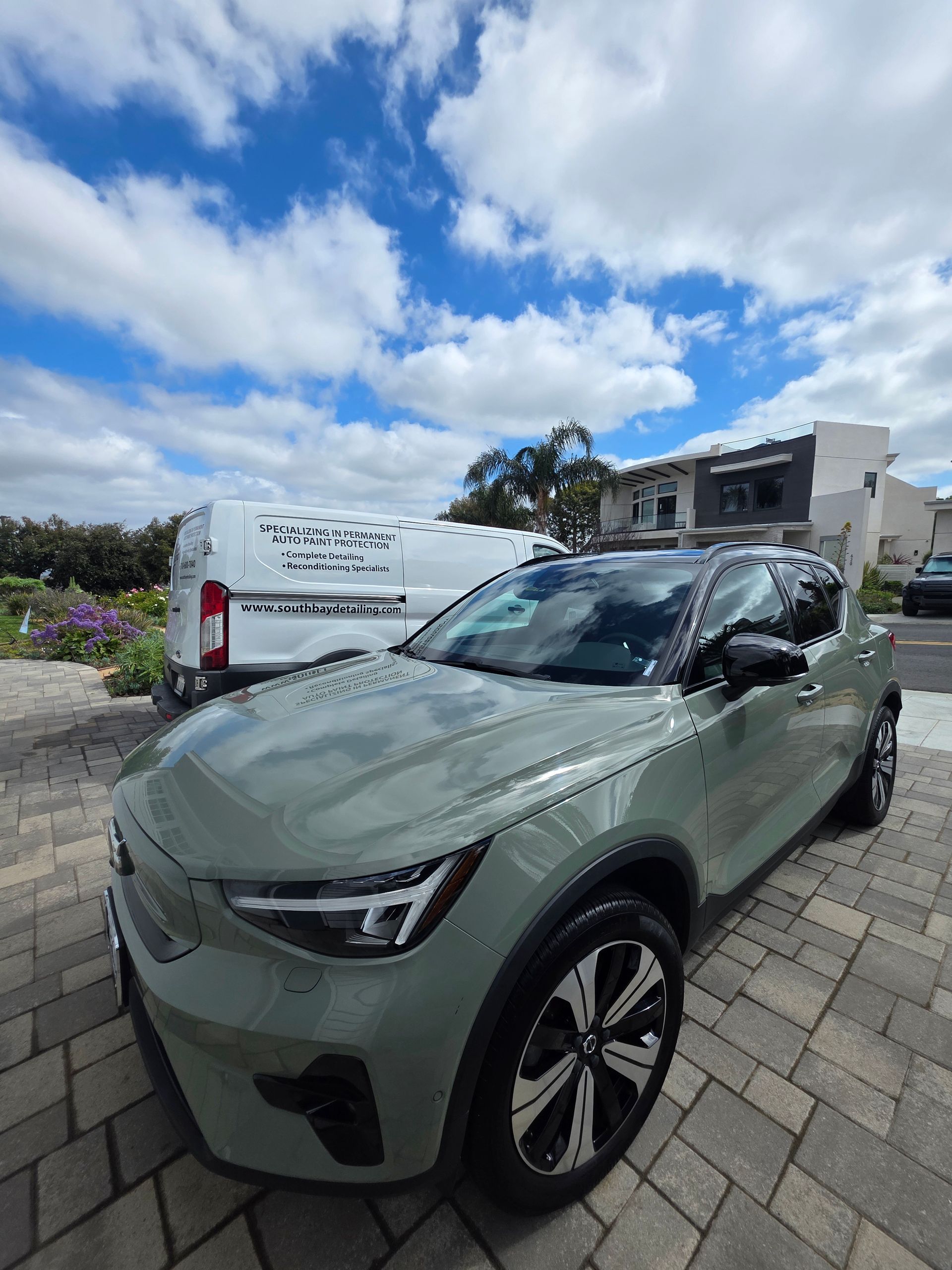 A light sage-green crossover vehicle parked on a paved driveway under a blue, cloudy sky.