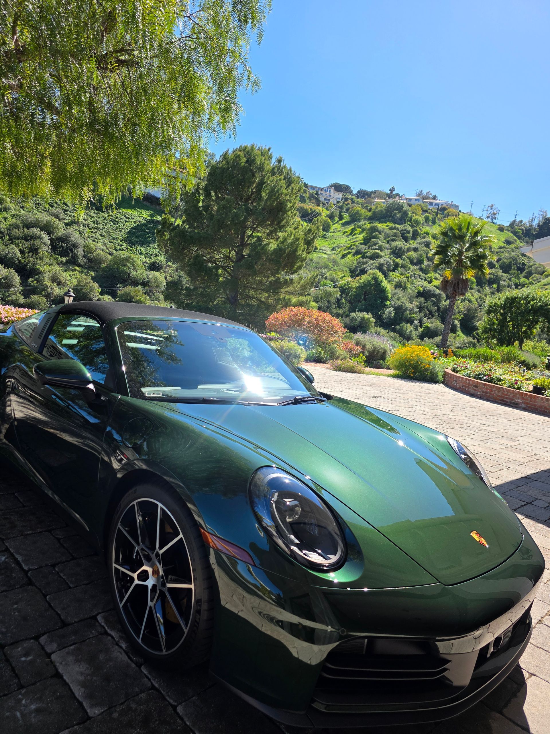 A dark green Porsche 911 parked on a paved driveway with a lush green hillside and blue sky in the background.