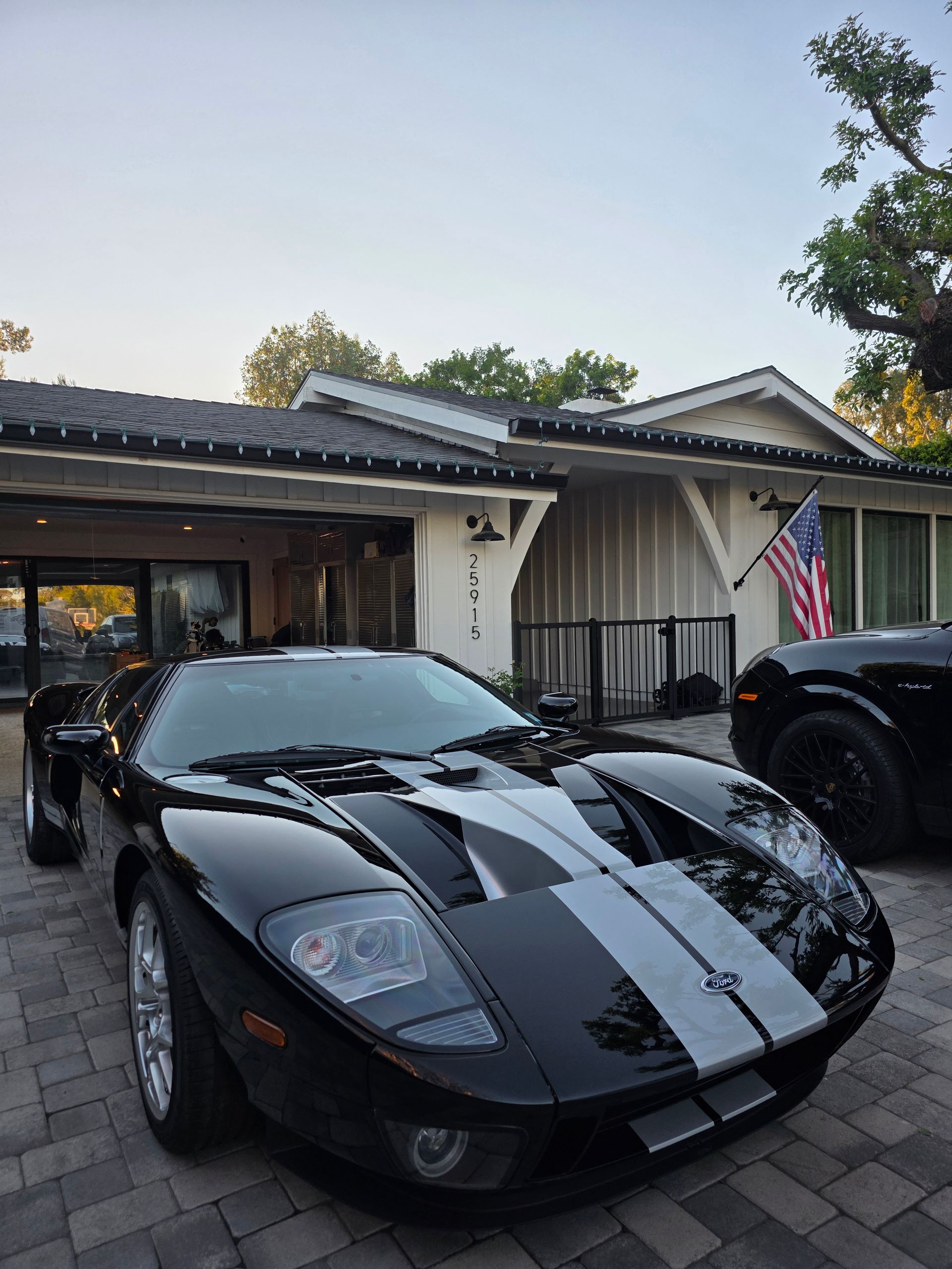 A black Ford GT with dual silver racing stripes parked on a brick driveway in front of a house.