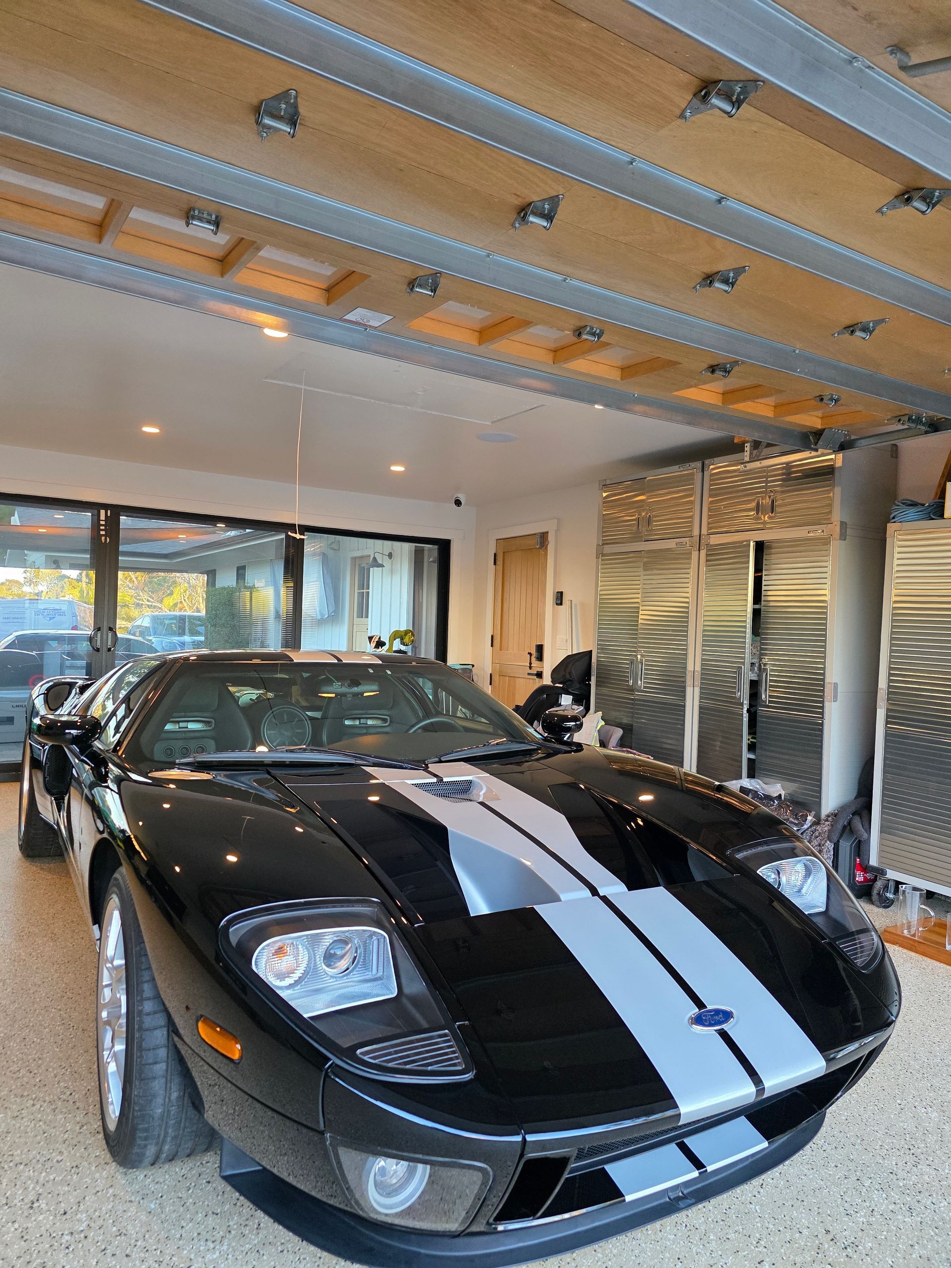A black Ford GT supercar with silver racing stripes parked inside a bright, modern garage with stainless steel cabinets.