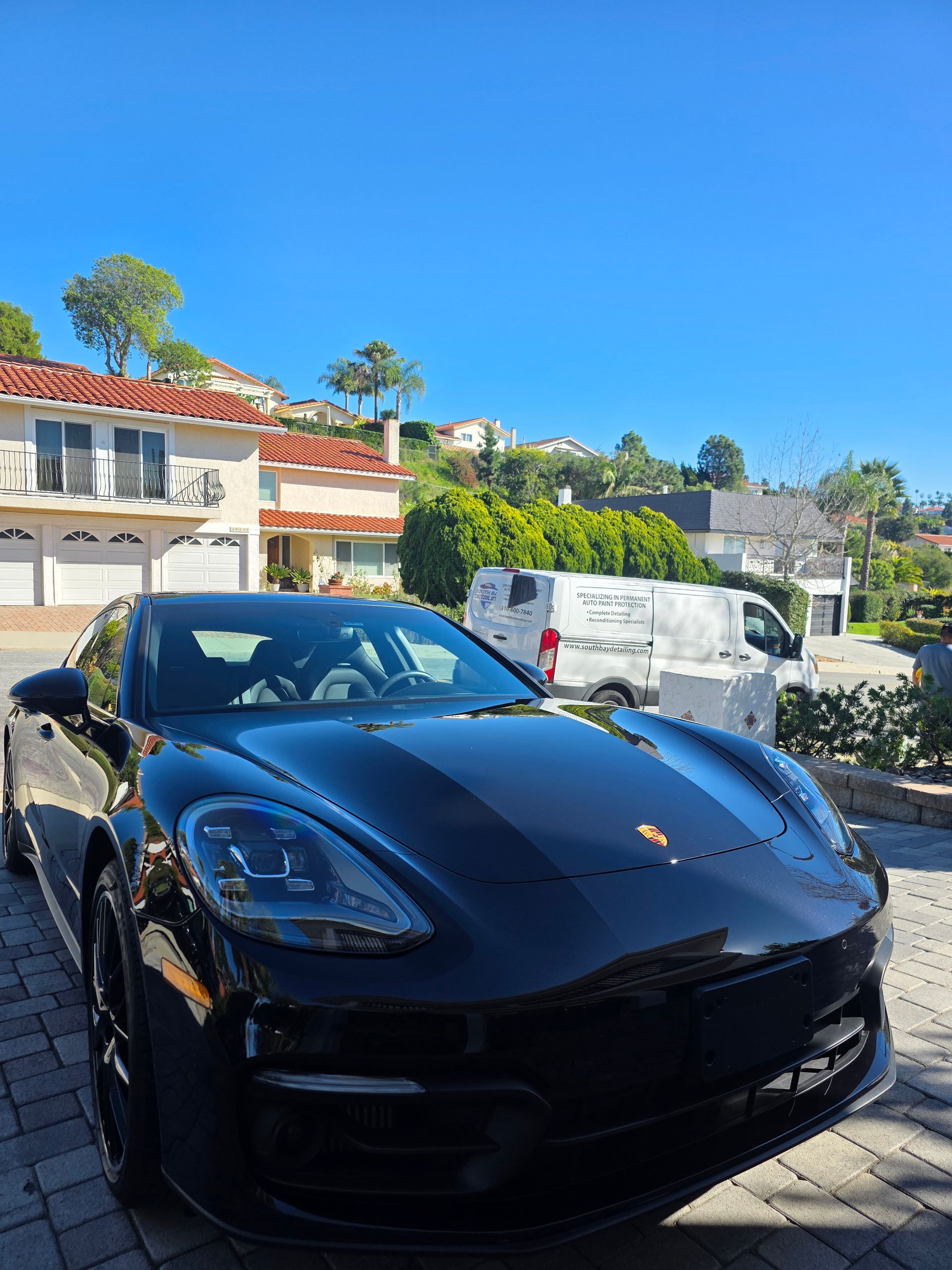 A sleek black Porsche parked on a paved driveway in front of a house on a sunny day.