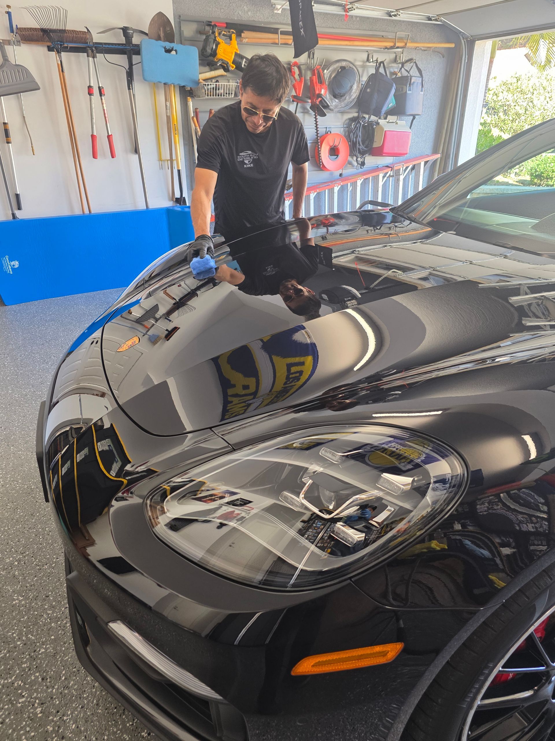 A person in a garage uses a blue sponge to wash the hood of a shiny black Porsche sports car.