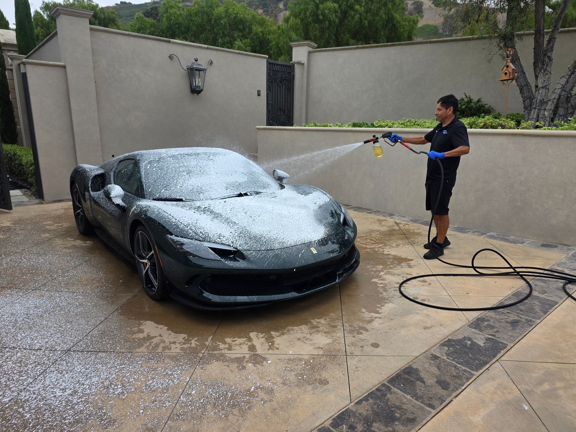 Person pressure-washing a black sports car in a driveway with soapy foam.