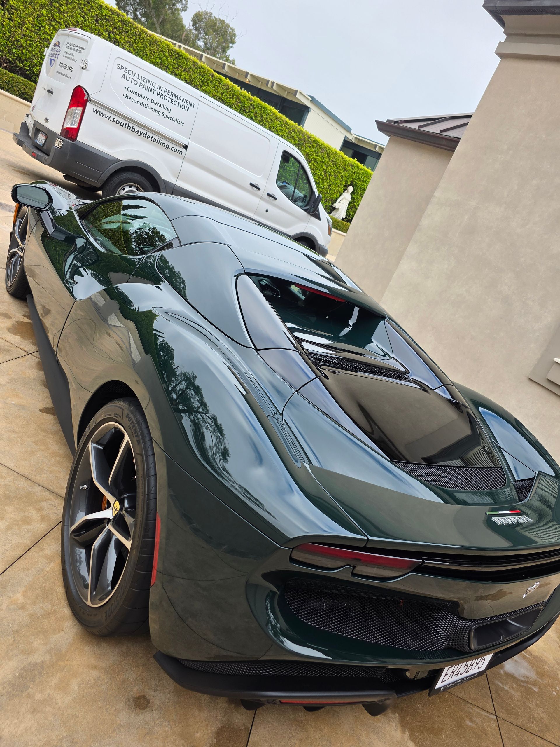Dark green sports car parked by a beige wall with a white van behind it.