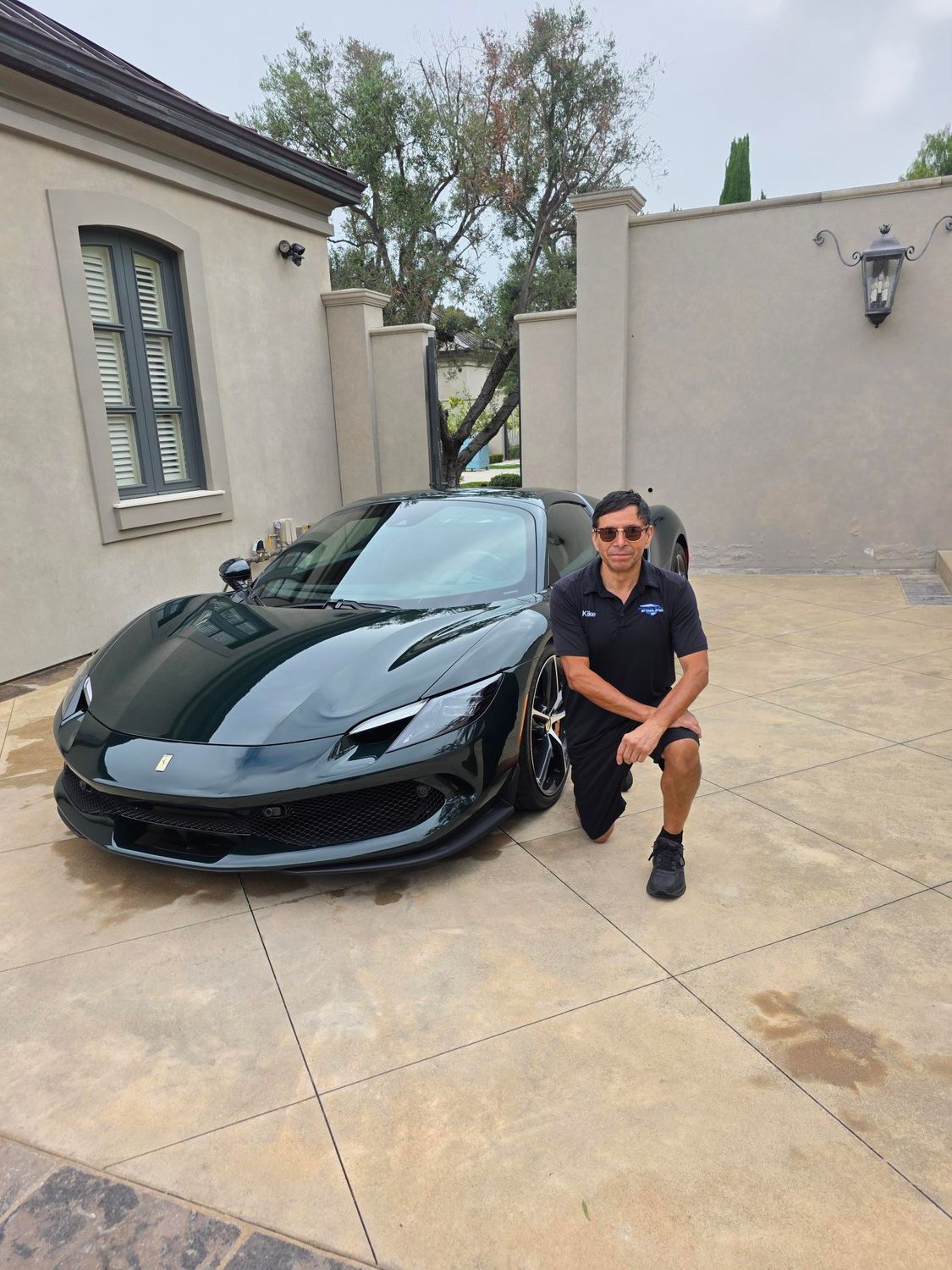 Man crouching beside a black sports car in a driveway outside a stucco house.