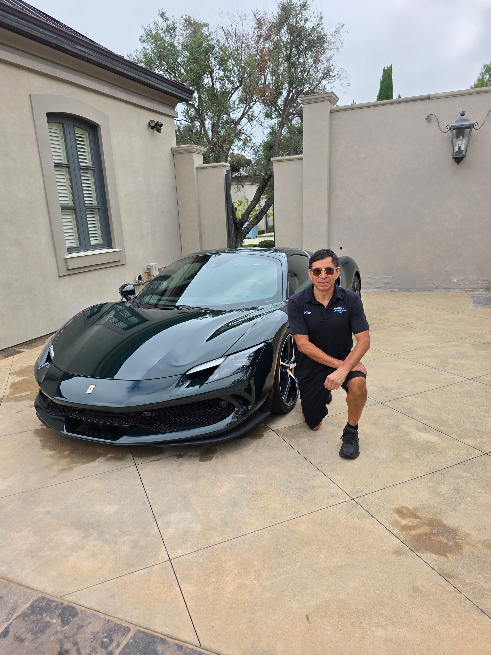 Man kneeling beside a black sports car in a driveway, with a house and wall in the background.