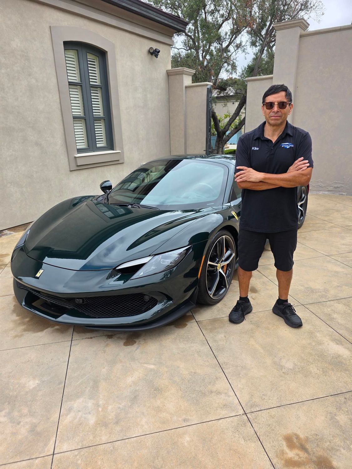 Man standing beside a black sports car in a driveway, arms crossed, wearing sunglasses.