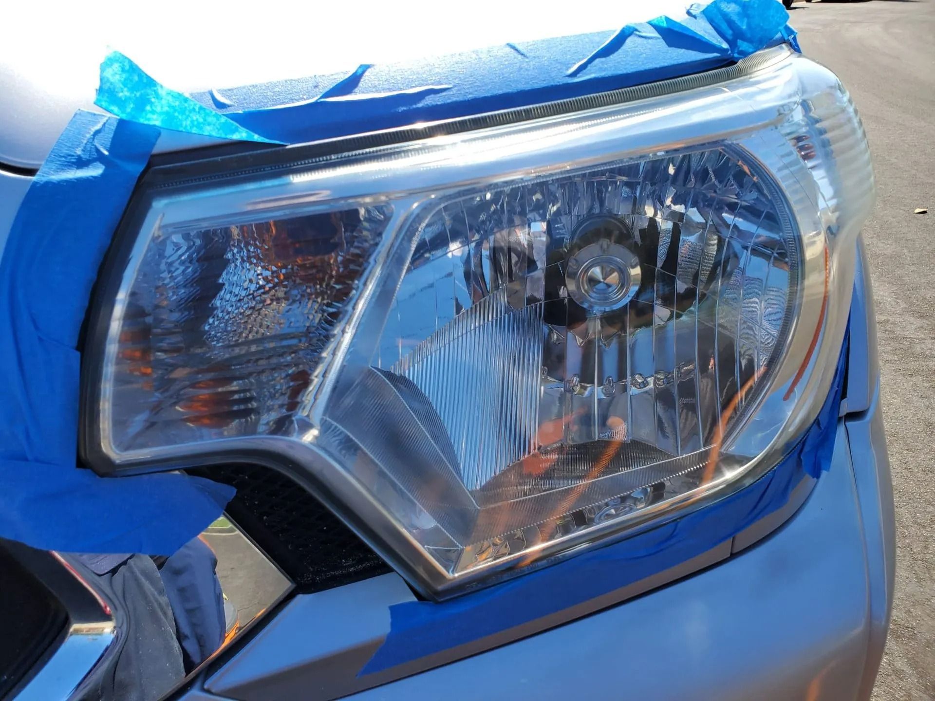 A silver car headlight masked with blue painter's tape, prepared for restoration or maintenance.