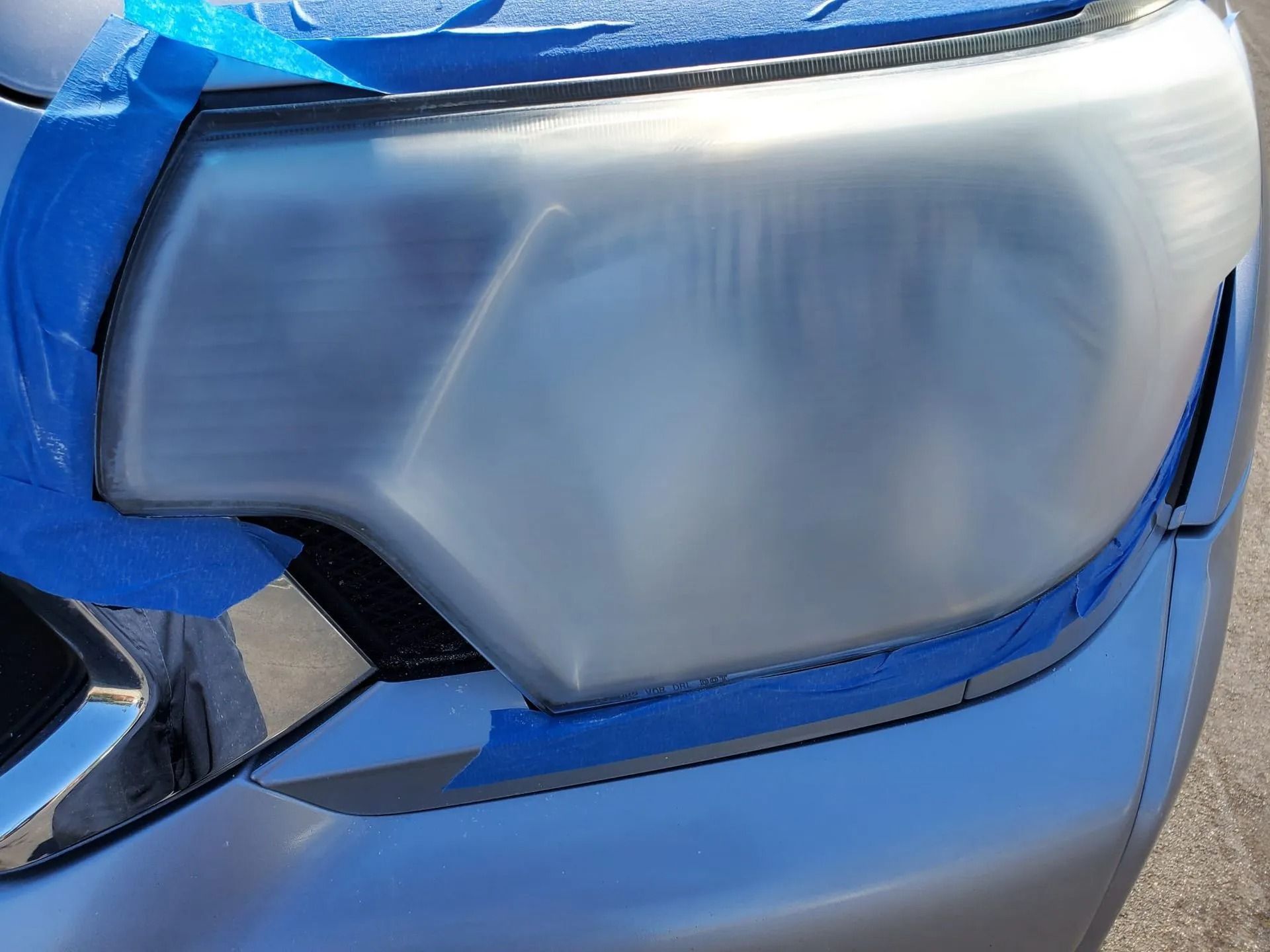 A car headlight covered in painter's tape, appearing sanded and clouded during a restoration process.