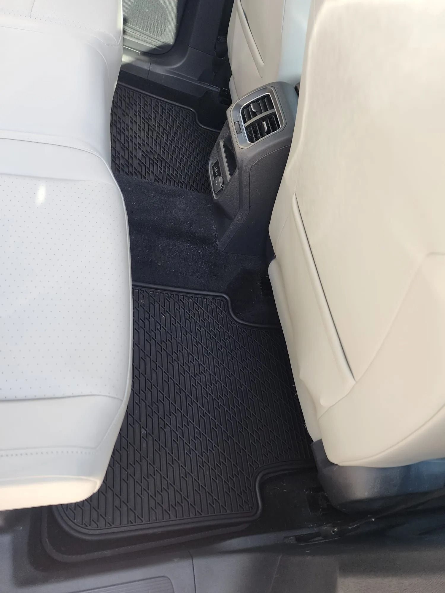 Interior view of a vehicle's back seat, showing black floor mats on dark carpet and the rear air conditioning console.
