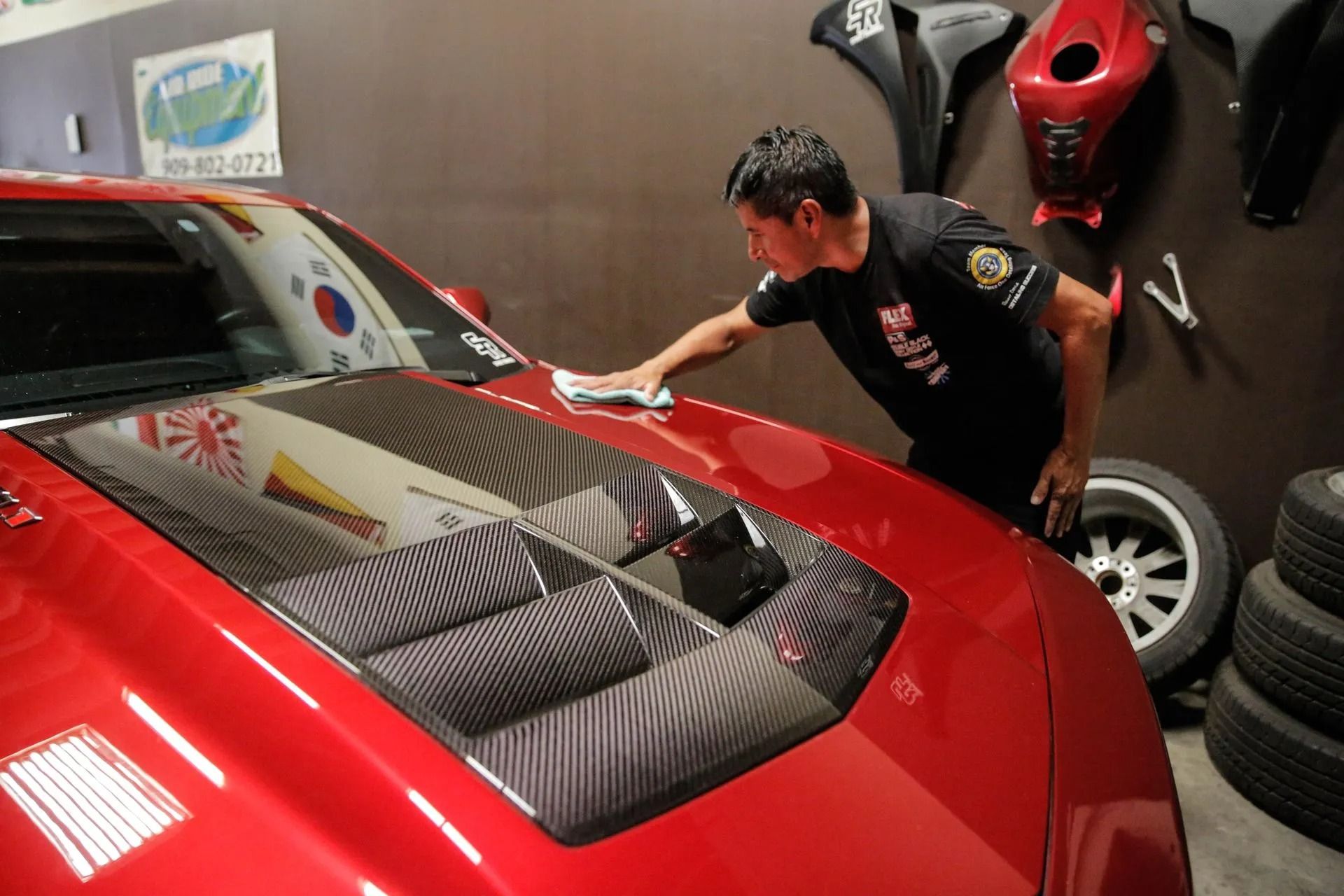A person polishing the carbon fiber hood of a red sports car in a garage with motorcycle parts on the wall.