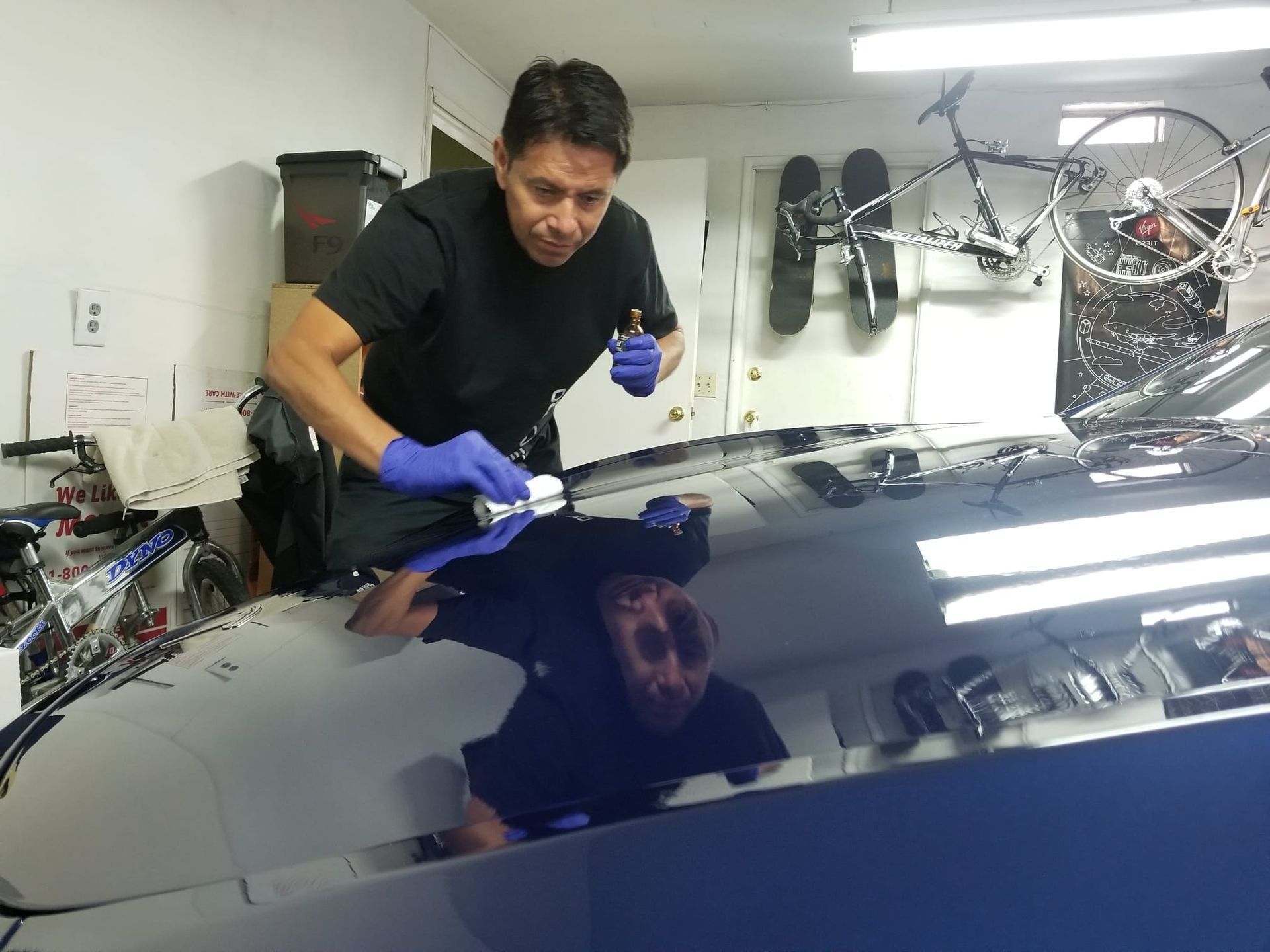 A person wearing blue nitrile gloves carefully applies detailing product to the shiny, dark hood of a car in a garage.