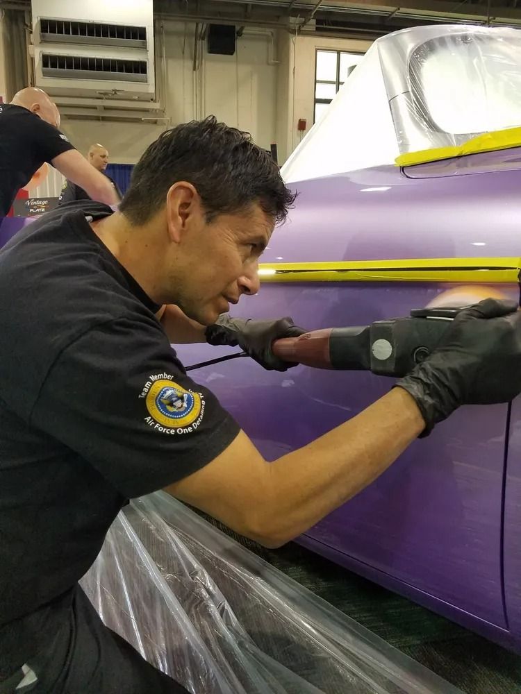 A technician in a black shirt and gloves uses a power buffer to polish the side of a light purple vehicle in a workshop.