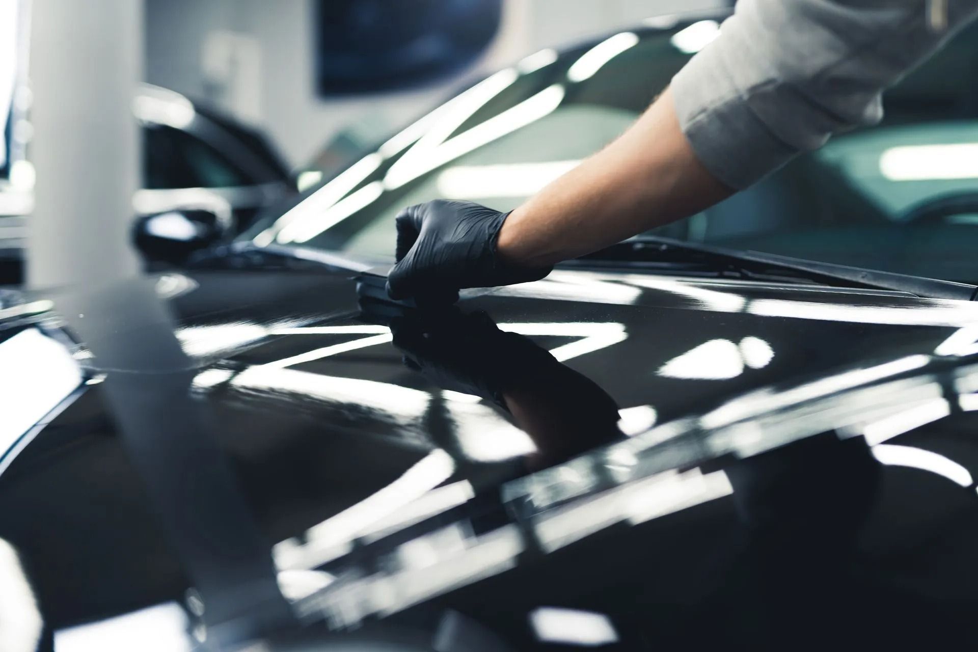 A person wearing a black glove applies a cleaning or polishing product to the glossy black hood of a car.