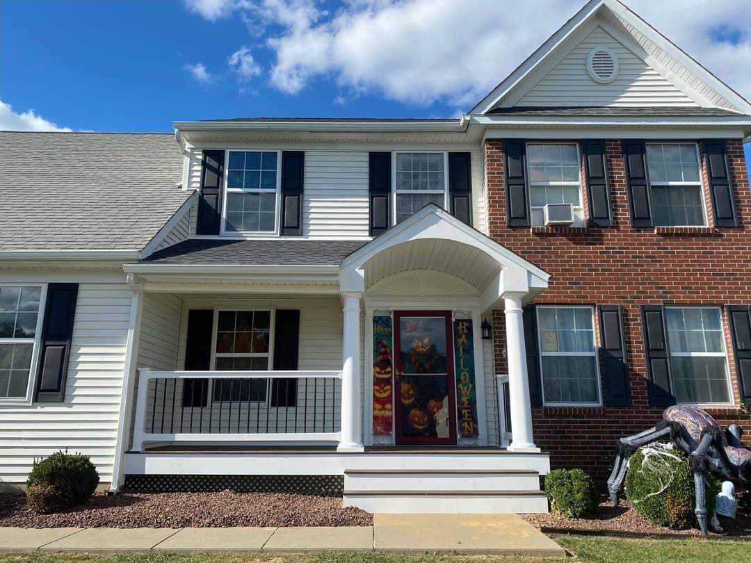 Two-story house with white siding and a brick section, black shutters, and a porch under a blue sky.