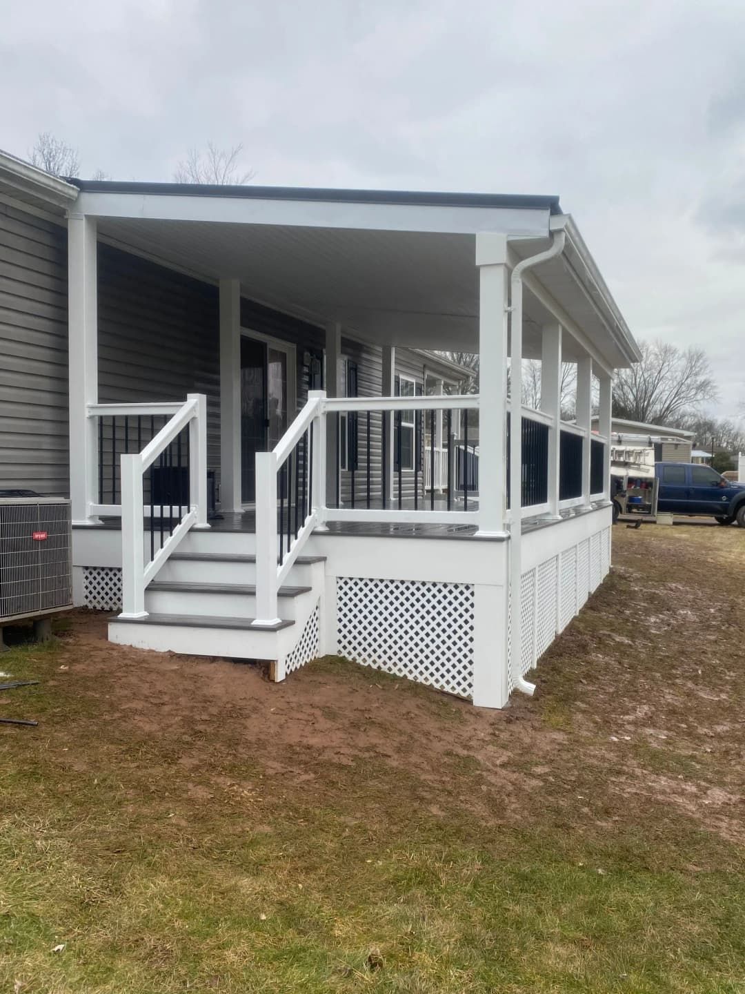 White-framed porch with stairs, black railings, and lattice skirting, attached to a gray house, set on brown grass.