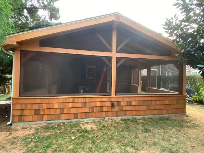 Wooden screened-in porch with cedar shake siding, built on a grassy area, in front of a dark-colored wall.