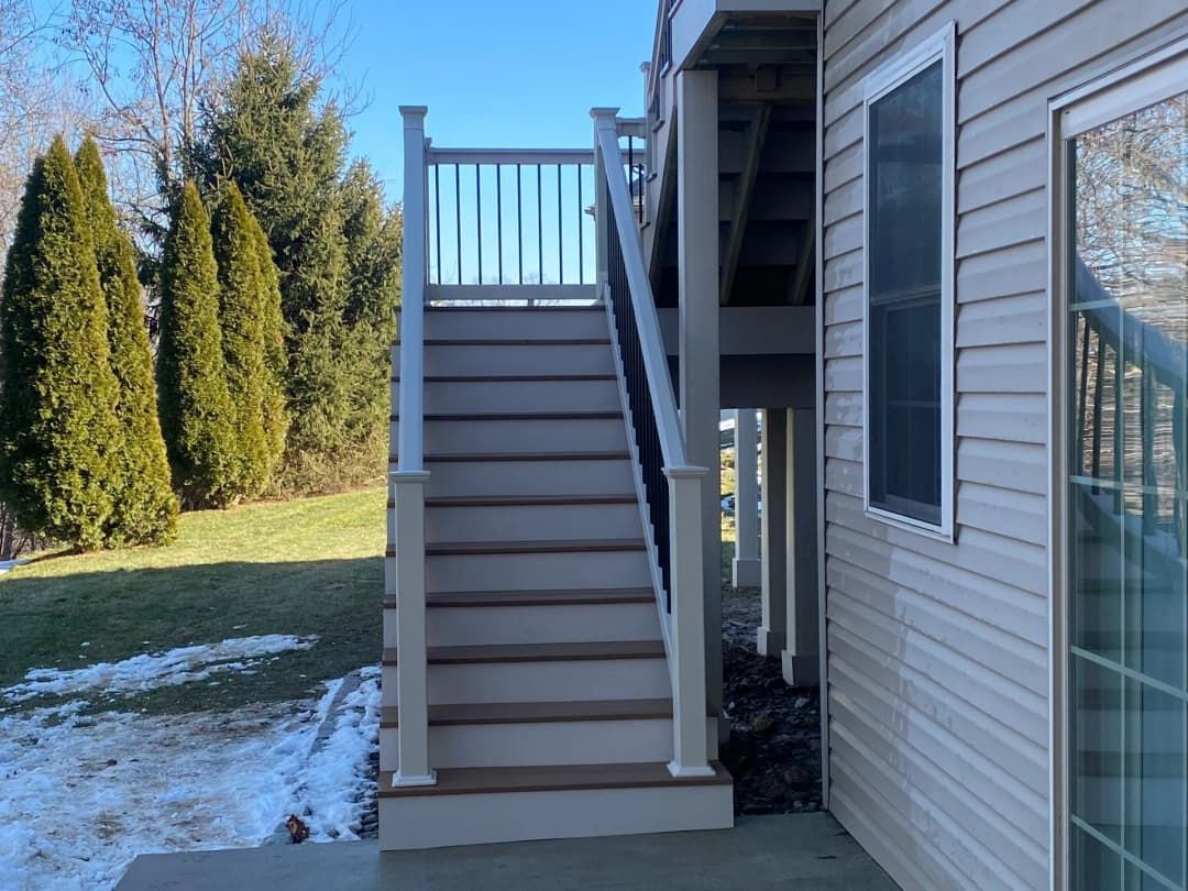 Outdoor staircase leading up to a deck, adjacent to a beige house, with snow on the ground and trees in the background.