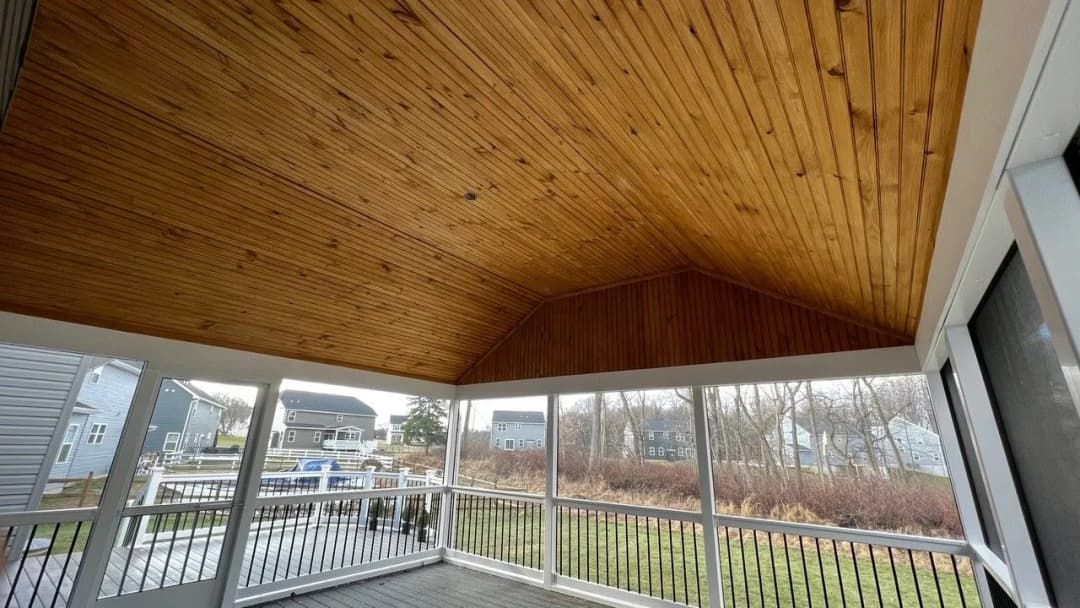 Screened porch with wooden ceiling, white railings, and a view of houses and water.
