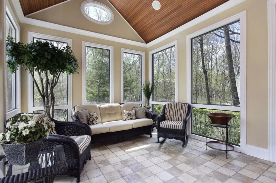 Sunroom with wicker furniture, windows overlooking trees, beige walls, and a tiled floor.
