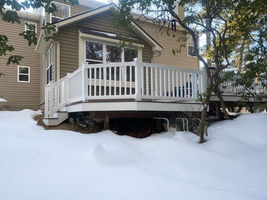Deck of a house covered in snow, with white railing and trees.