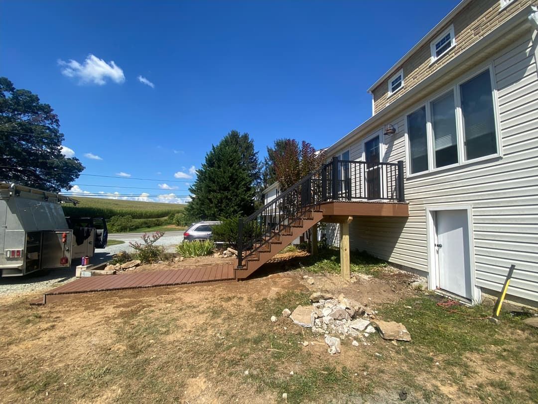 Backyard with a brown deck, stairs, black railing, and a white house under a blue sky.