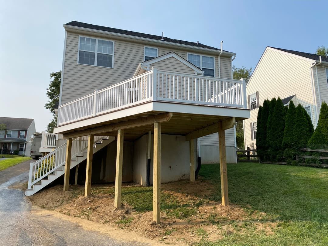 A raised wooden deck attached to a two-story house, with white railings and support posts, accessible by stairs.