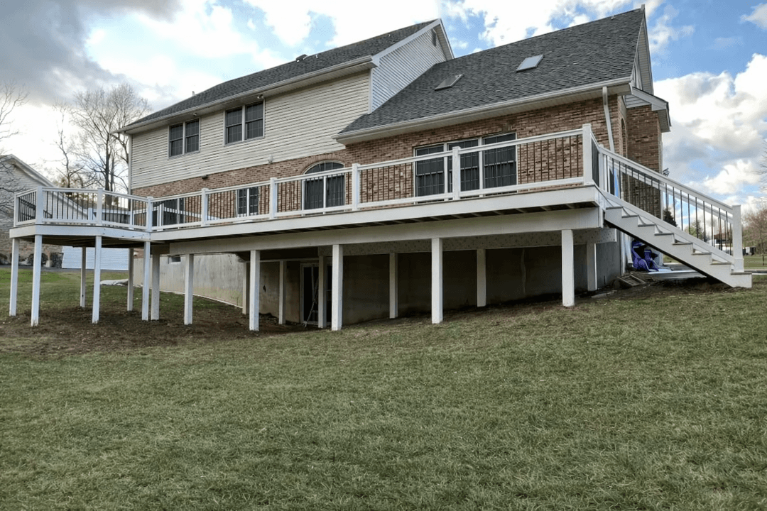 Two-story house with white deck and stairs, brick and siding exterior, on a grassy lawn.