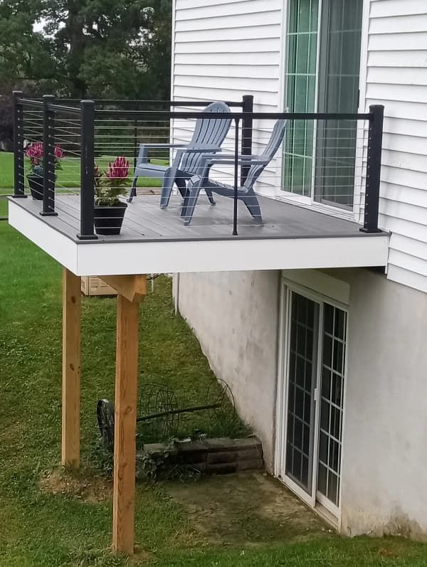 Deck with black railing, gray floorboards, and two blue chairs. Supported by wooden posts, next to a white house with a basement door.