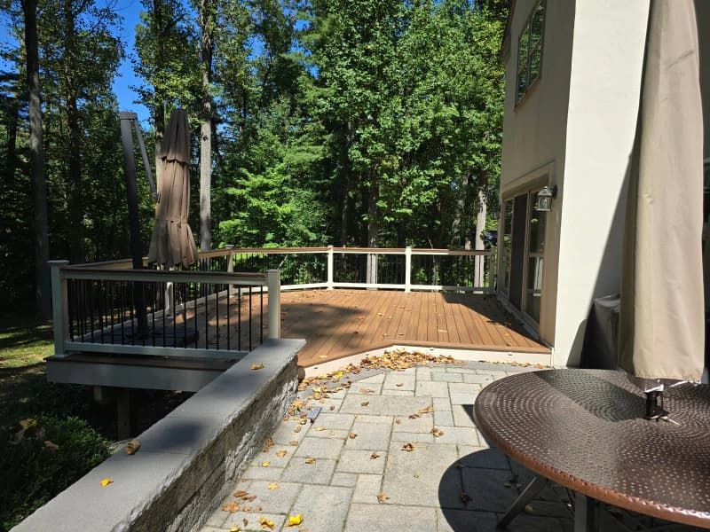 Backyard patio with a deck, trees, and a house on a sunny day. Brown and gray hues.