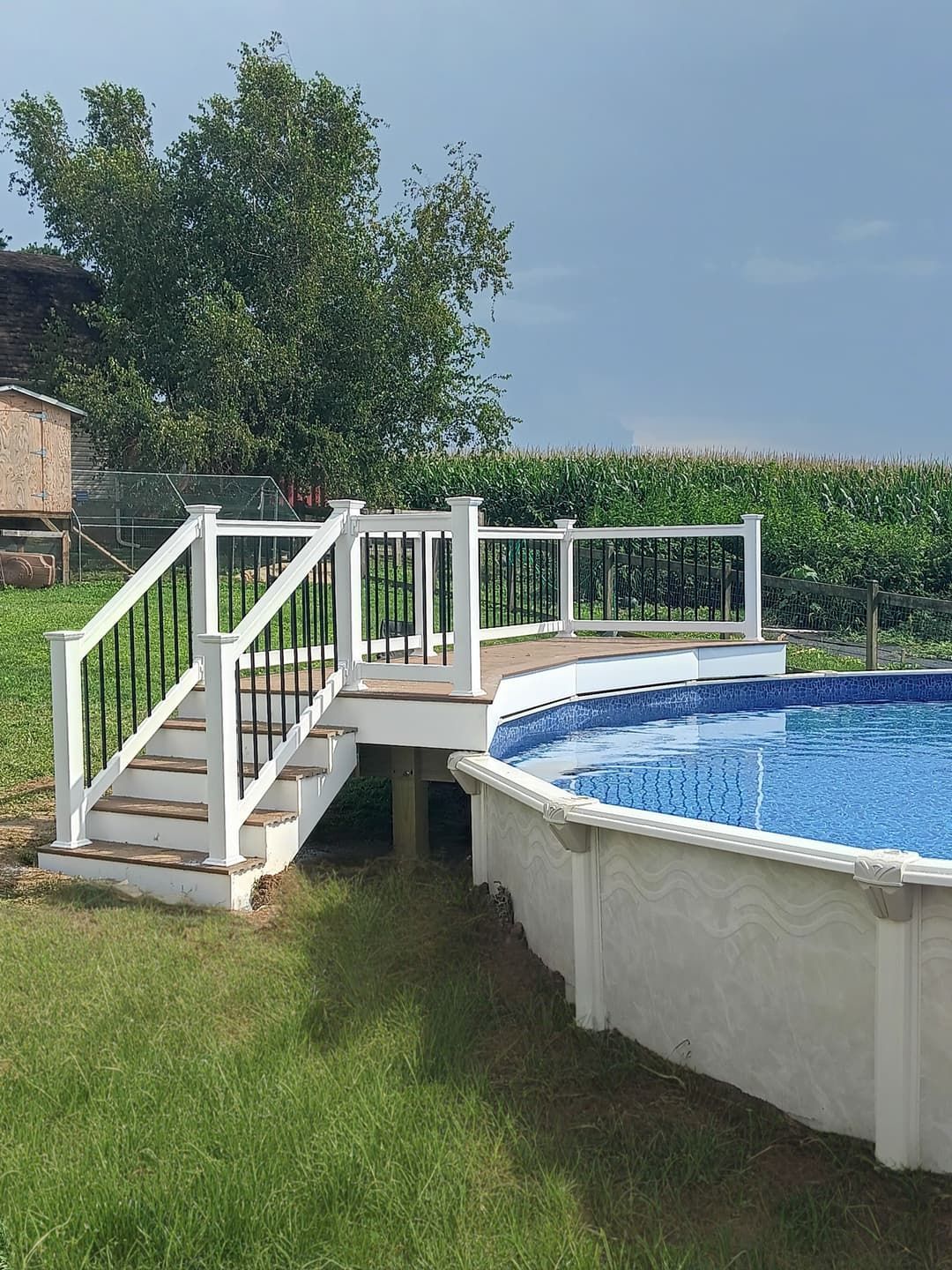 White deck with black railing and steps next to an above-ground pool, in a grassy backyard.